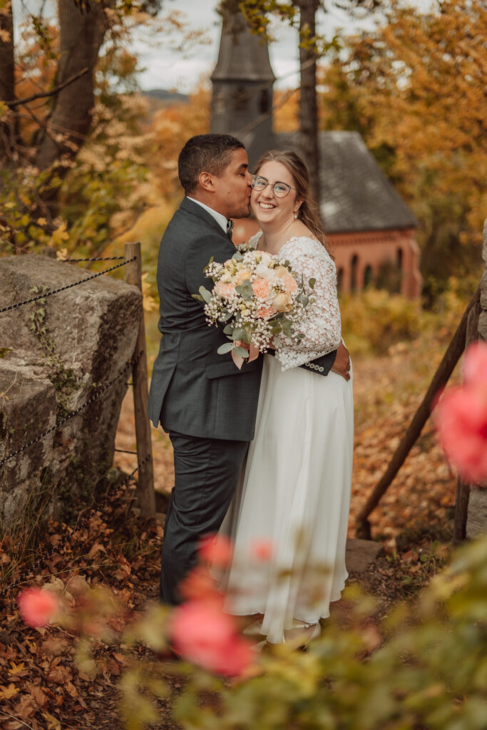 Bräutigam küsst die Braut mit Brautstrauß auf die Wange vor herbstlicher Kulisse mit Kapelle, im Vordergrund rosa Blüten