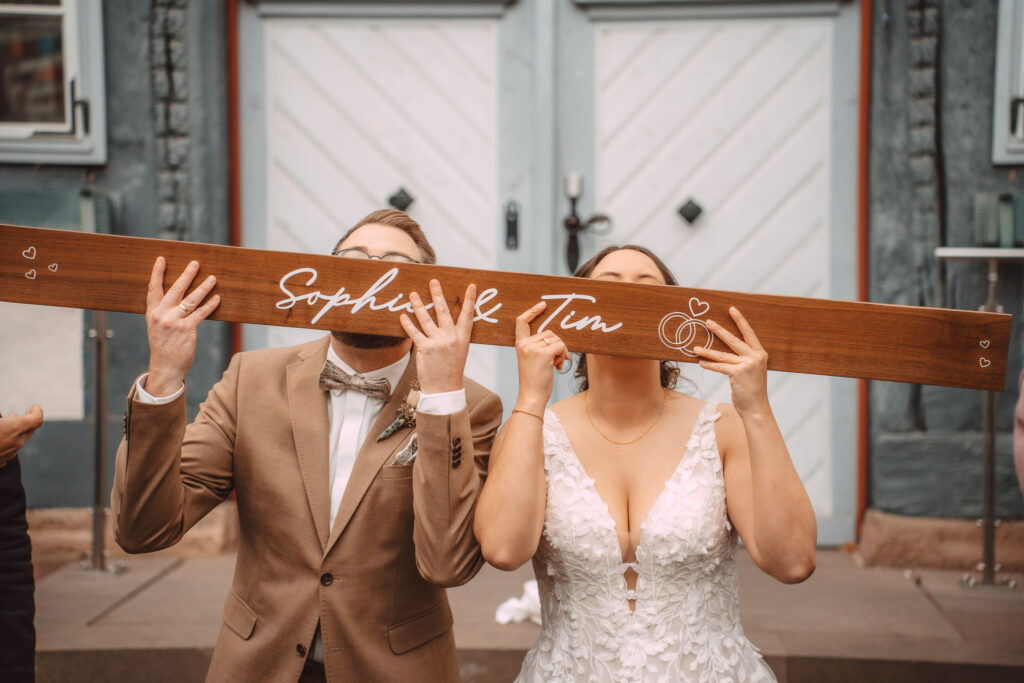 Brautpaar hält ein Holzschild mit dem Schriftzug Sophie und Tim vor den Gesichtern, vor blauer Altstadttür
