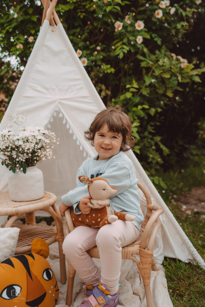 Lächelndes Kleinkind mit Plüsch-Reh auf einem Rattanstuhl vor einem weißen Tipi im Garten