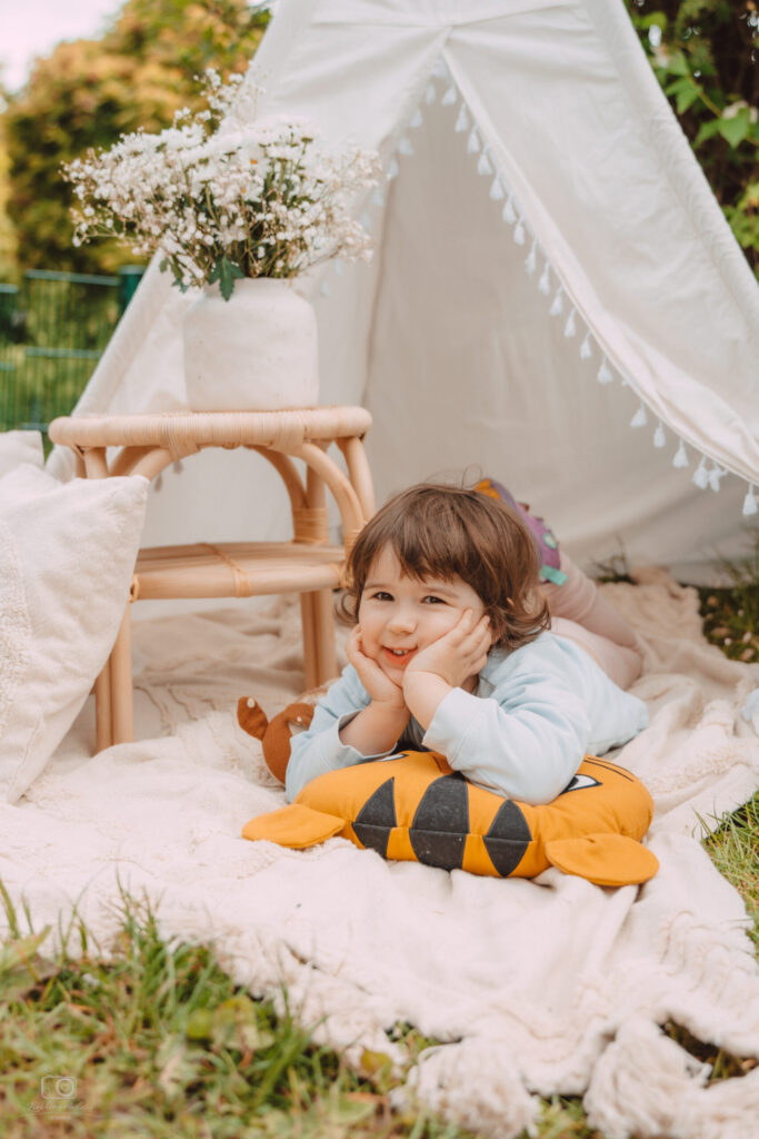 Kleinkind mit dunklen Haaren liegt lächelnd mit aufgestütztem Kopf auf einer hellen Decke vor einem Tipi-Zelt – Outdoor-Kinderporträt