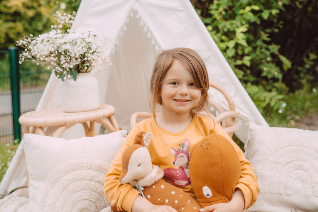 Lächelndes Mädchen im gelben Reh-Shirt sitzt mit Kuscheltieren vor einem weißen Tipi-Zelt im Garten – Kinderfotografie im Freien