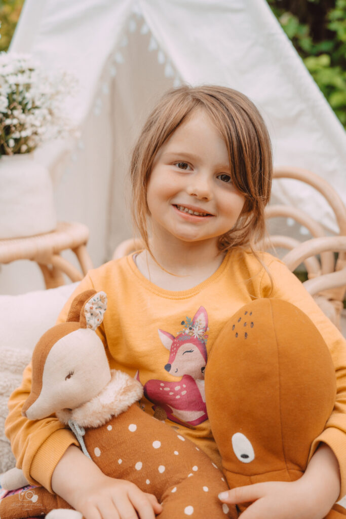 Lächelndes Mädchen mit hellbraunen Haaren hält zwei Reh-Kuscheltiere vor einem Tipi-Zelt – Kinderporträt im Freien